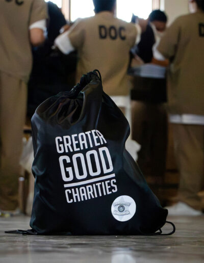 A close up of a black drawstring bag that has "Greater Good Charities" written on it. In the background are three individuals in custody.