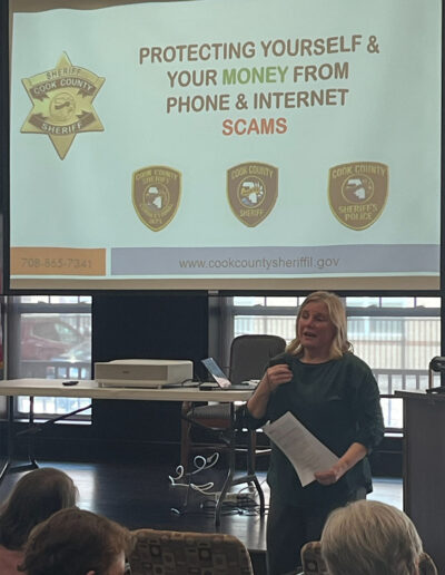 A woman addresses an audience. Behind her is a projector screen that reads: "Protecting yourself and your money from phone and internet scams."