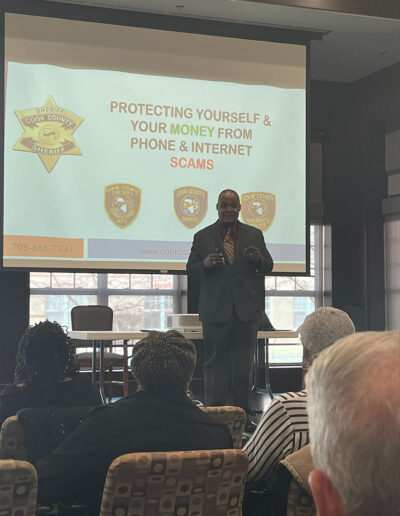 A man addresses an audience. Behind her is a projector screen that reads: "Protecting yourself and your money from phone and internet scams."