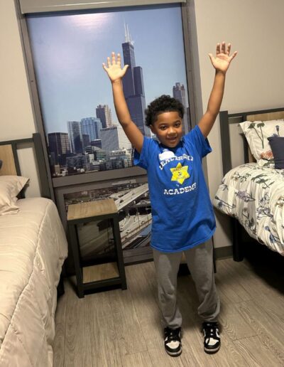 A young boy poses in front of a window with both arms raised above his head