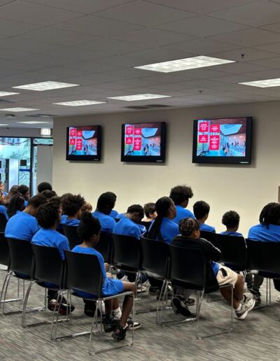 A woman addresses a group of children. There are TV screens displaying a presentation, but the text is unclear.